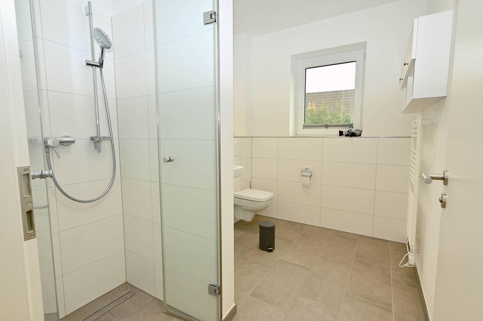 Bad mit Dusche, Toilette und Fenster. Fliesenboden und weiße Wände.