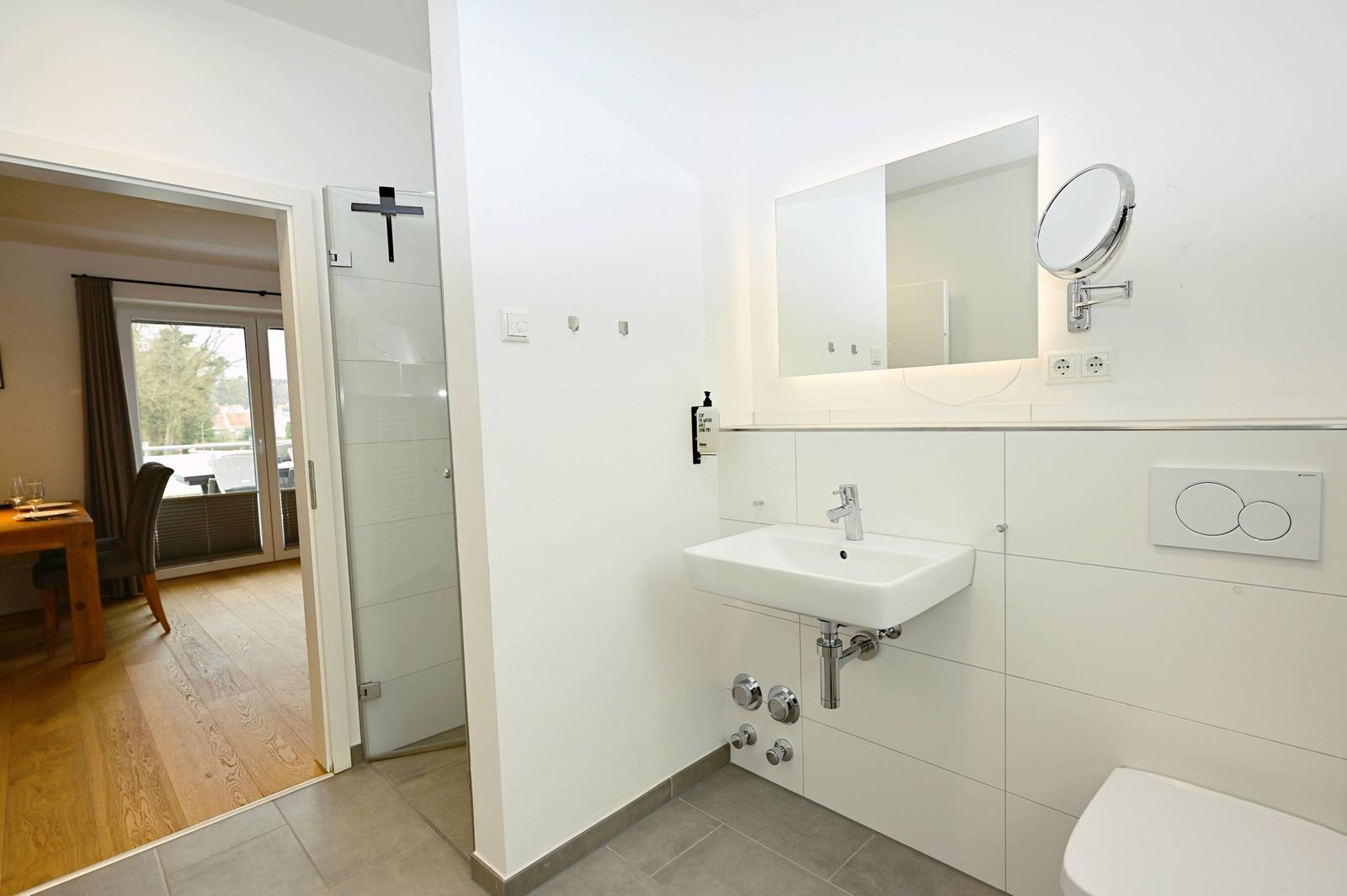 Badezimmer mit Waschbecken, Spiegel und Toilette. Durchgang zu Wohnbereich mit Holzboden.