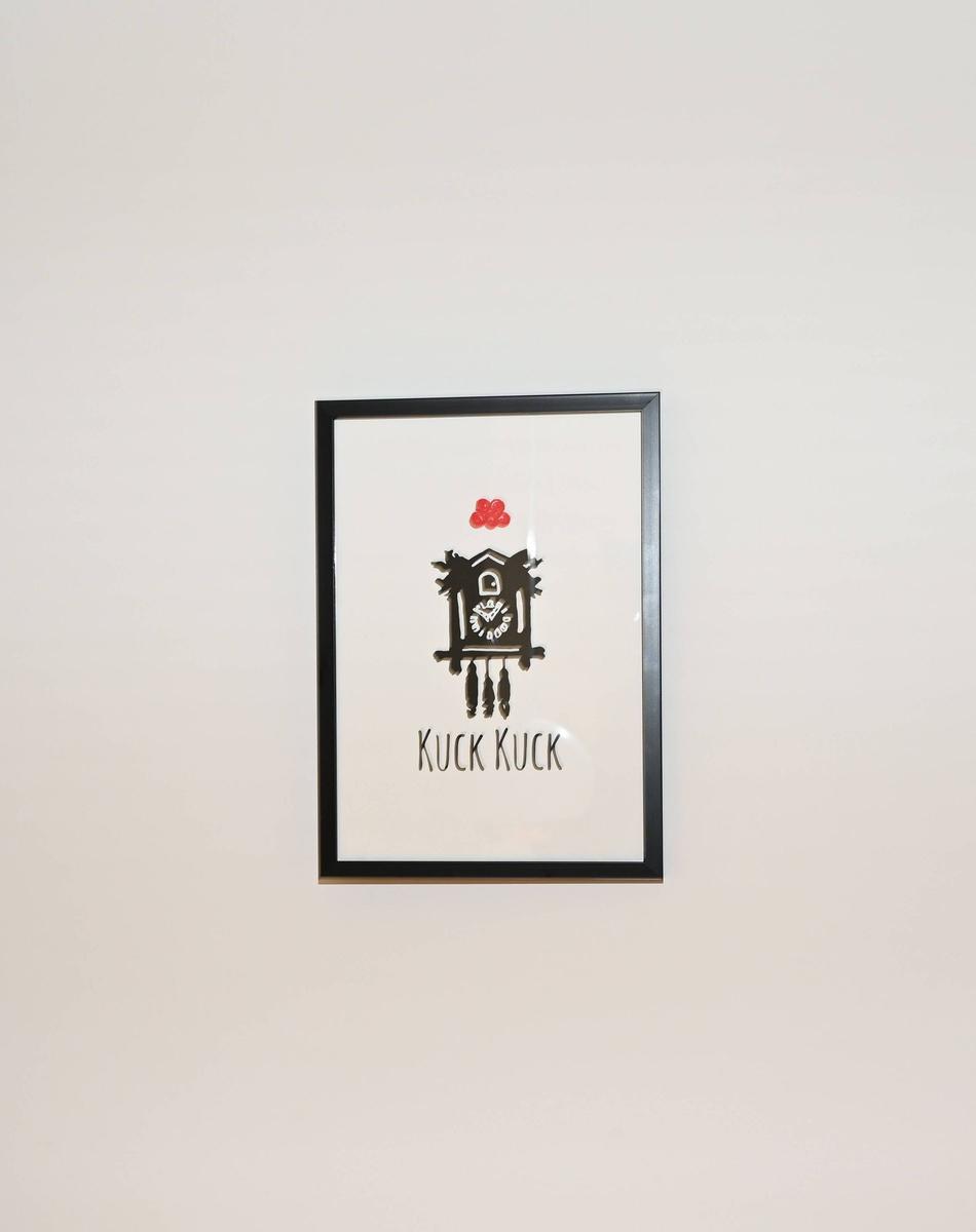 Ein gerahmtes Bild einer schwarzen Kuckucksuhr mit rotem Dach und dem Text Kuck Kuck.