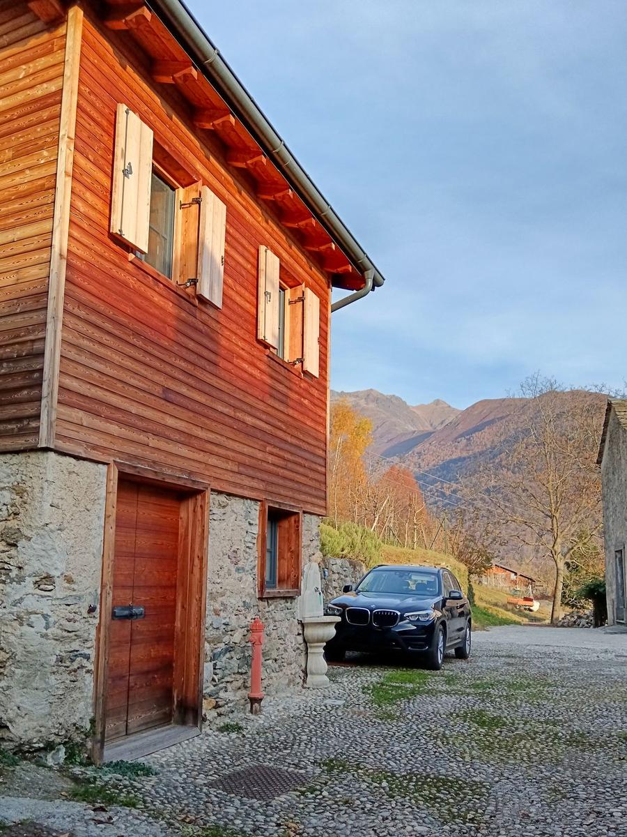 Holzhaus mit Steinbasis, schwarzes Auto auf Kiesweg, Berge im Hintergrund