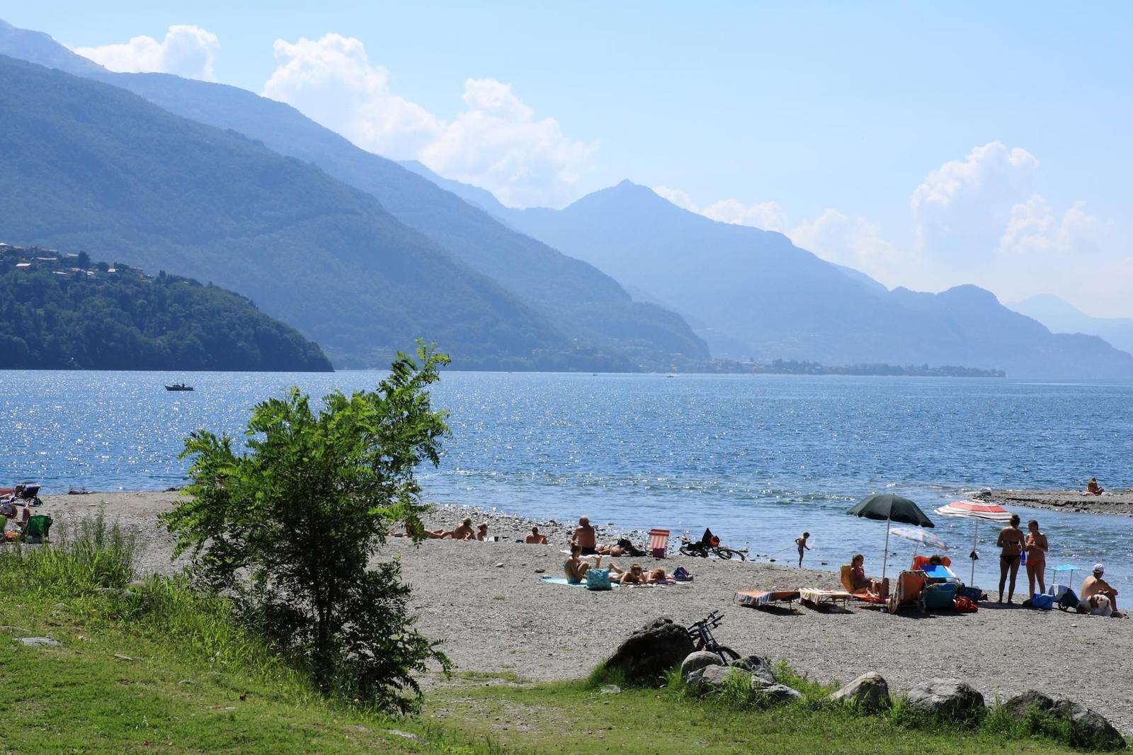 Strand mit Steinen und Bäumen am See mit Bergen im Hintergrund.