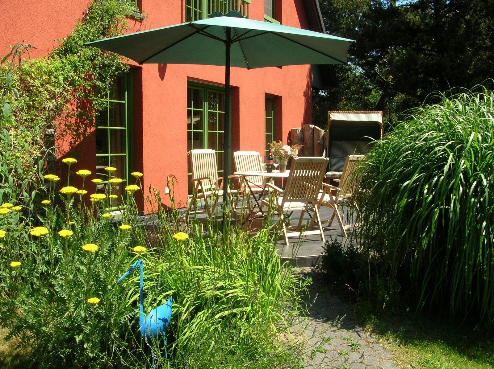 Terrasse mit Tisch, Stühlen und Sonnenschirm vor rotem Haus mit grünen Fenstern.