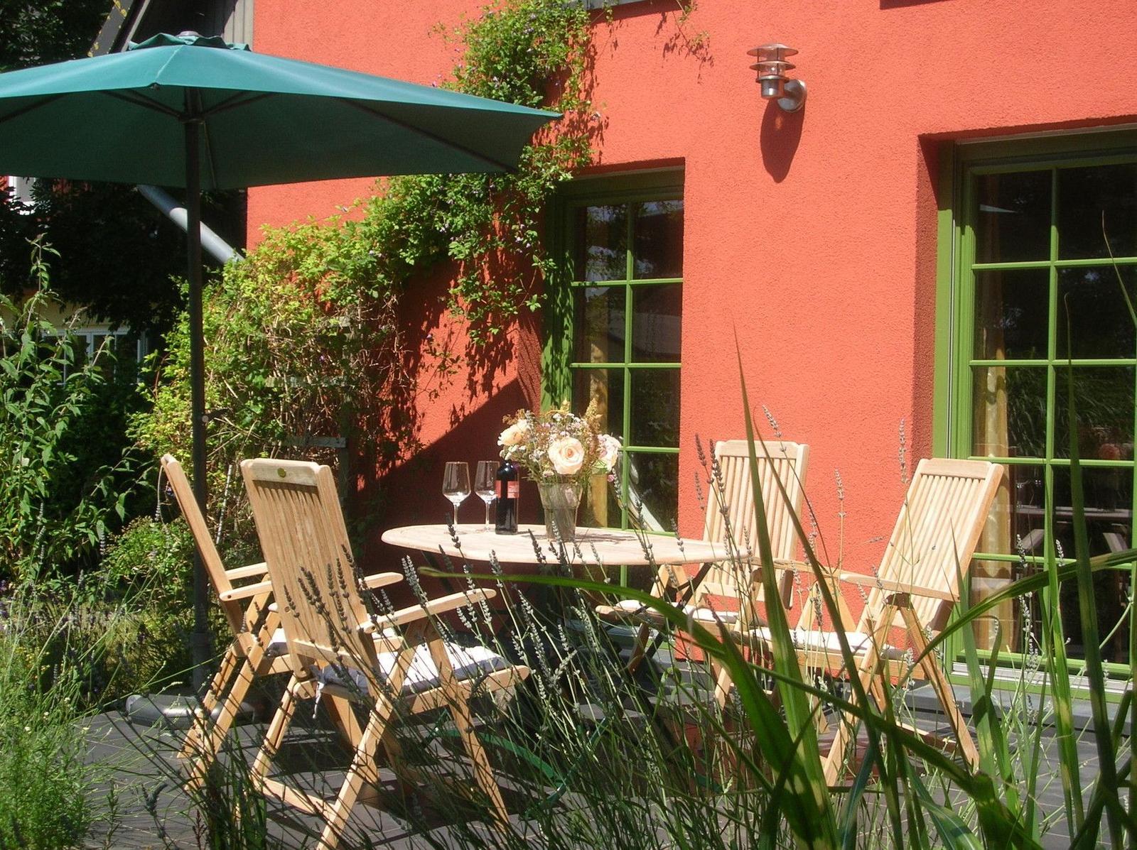 Terrasse mit Tisch, Stühlen und Sonnenschirm vor rotem Haus mit grünen Fenstern.