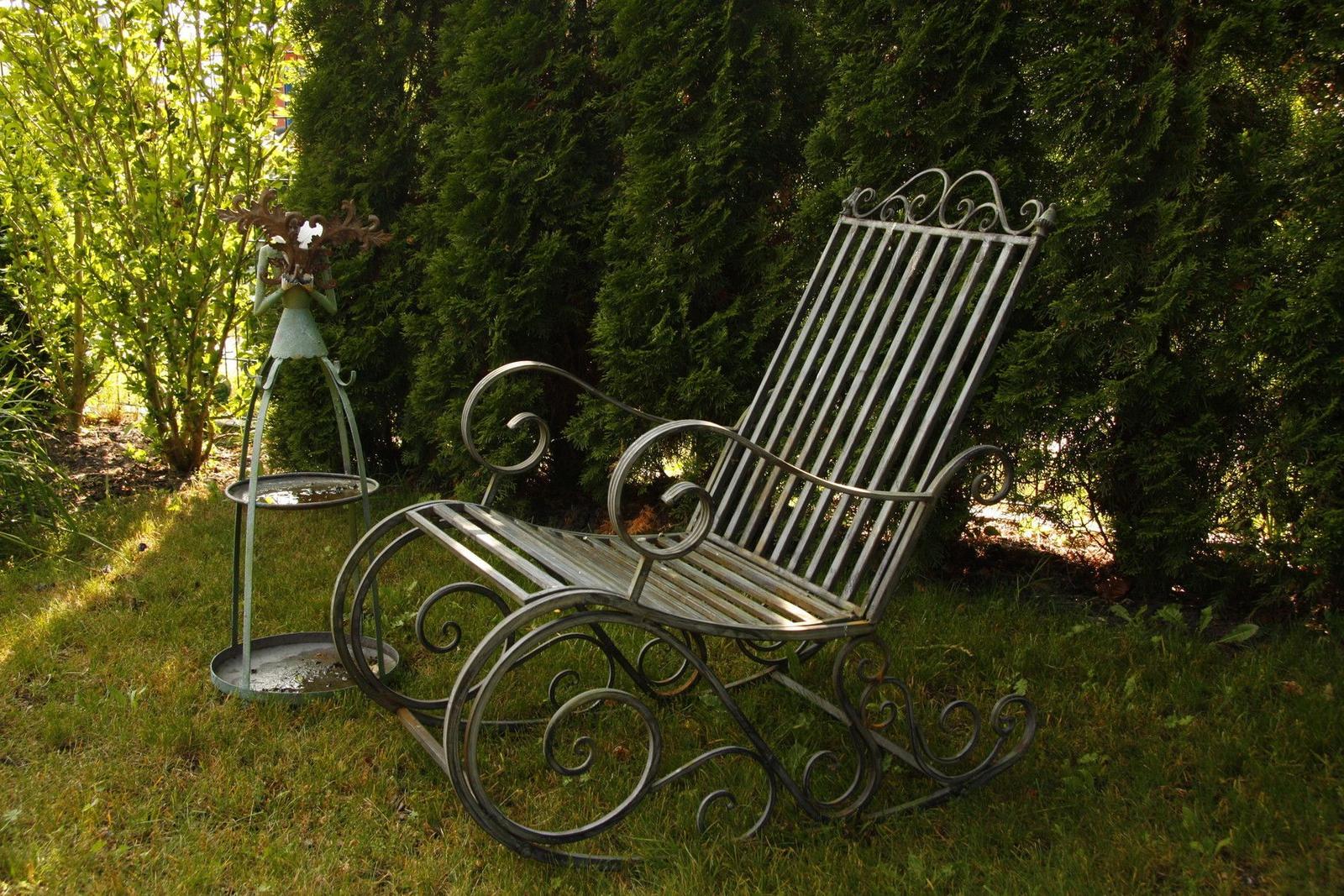 Schwungvolle Metall-Schaukelstuhl im Garten neben einer Pflanzentonne.