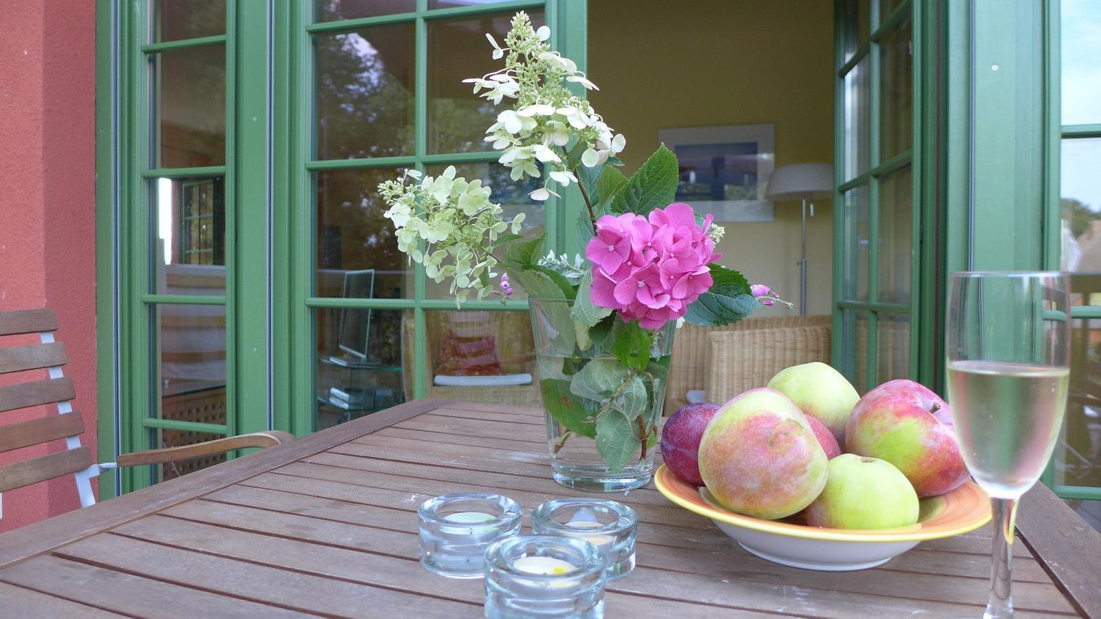 Holztisch mit Blumen, Obst und Wein vor grünen Fenstern.