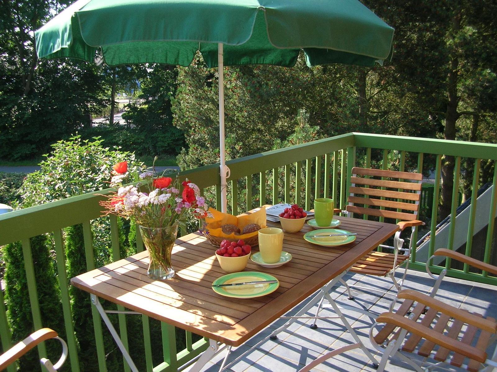 Terrasse mit Tisch, Stühlen und grünem Sonnenschirm. Frühstückstablett mit Obst und Blumen.