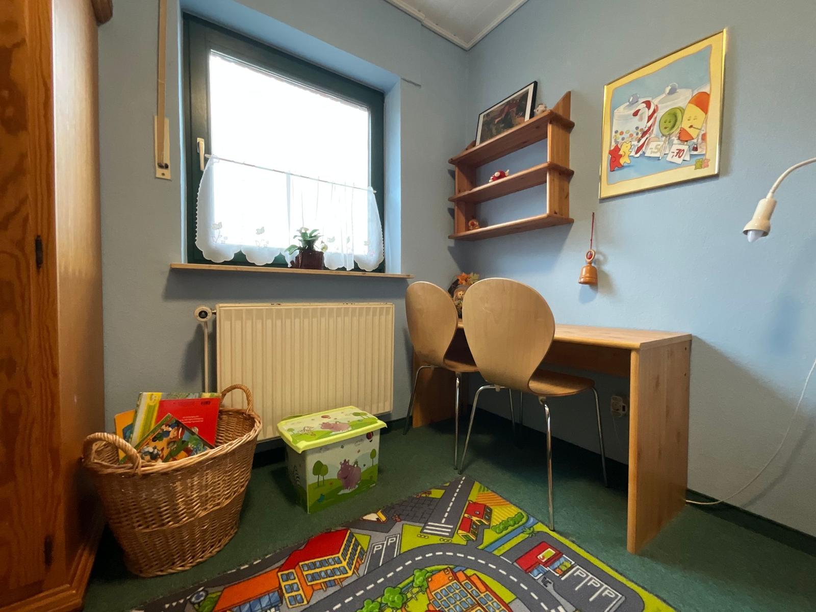 Ein Kinderzimmer mit Schreibtisch, Stühlen, Spielteppich und Bücherkorb.