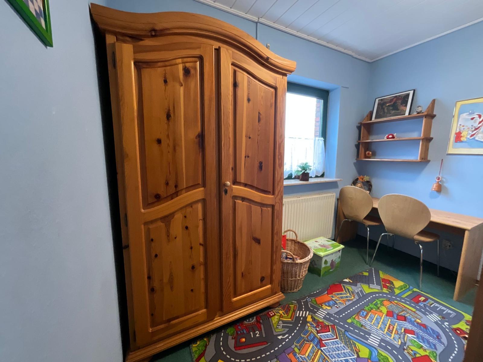Ein Zimmer mit Holzschrank, Schreibtisch, Stühlen und Spielteppich.