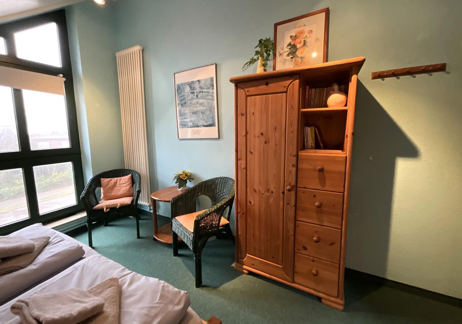 Zimmer mit Bett, Holzschrank, zwei Stühlen und Fenster.