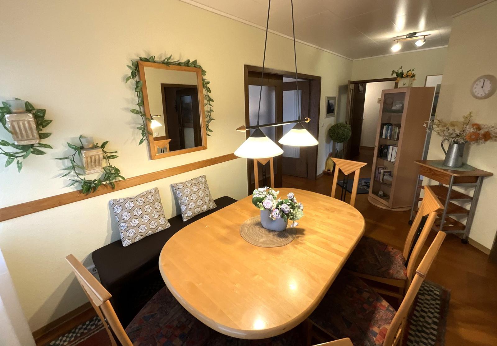 Esszimmer mit Holztisch, Stühlen und Wandspiegel. Bücherregal und Blumenvase sind sichtbar.