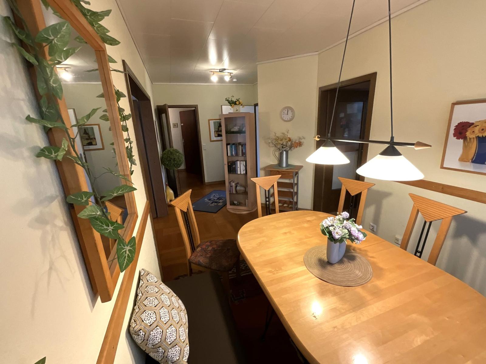 Essbereich mit Holztisch, Stühlen und Deckenleuchte. Im Hintergrund Bücherregal und Flur.