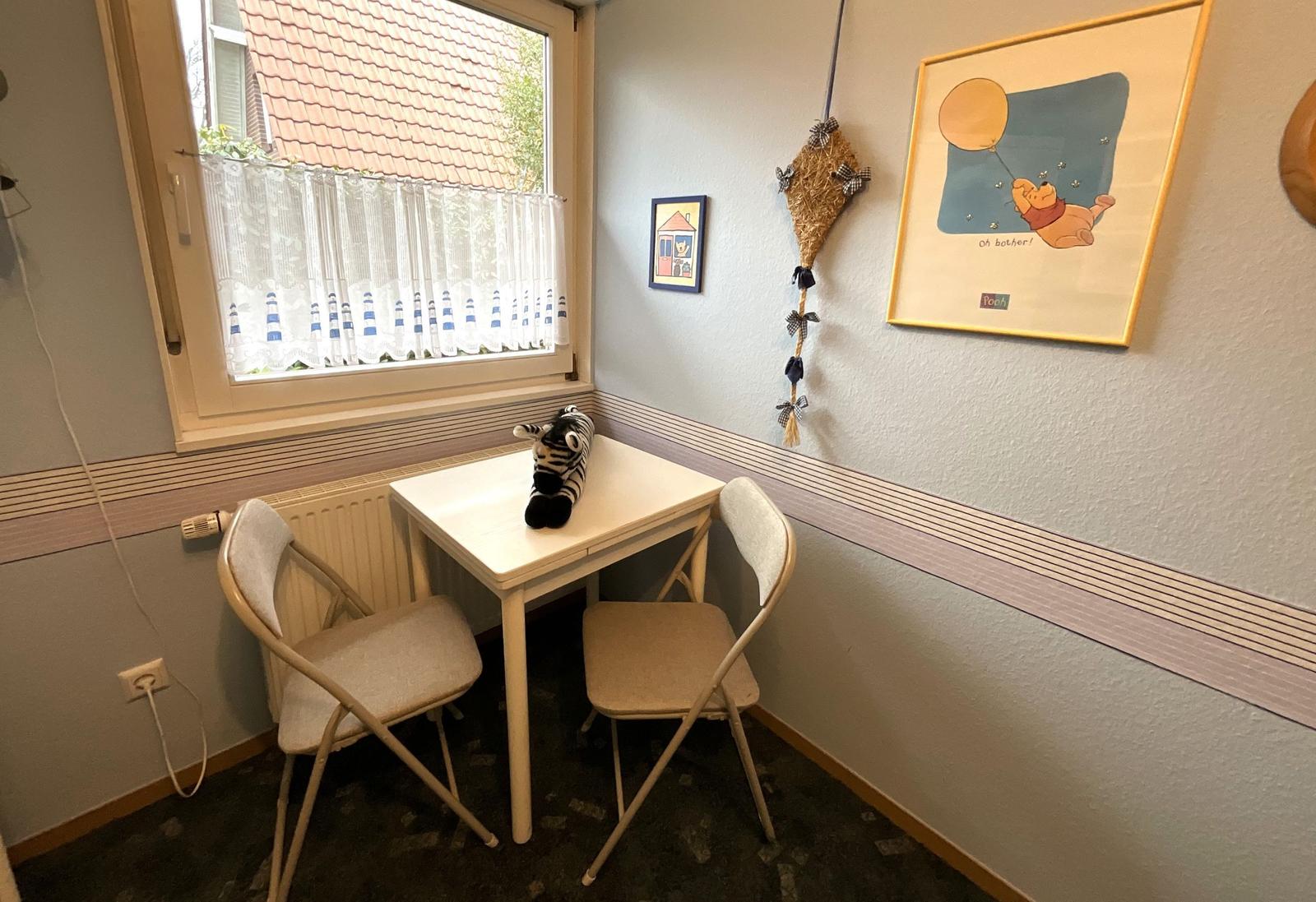 Kleiner Raum mit Tisch, zwei Stühlen und Fenster. Wandbild und Spielzeug sind sichtbar.