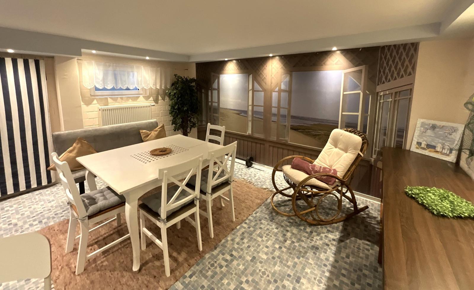 Esszimmer mit Tisch, Stühlen und Sessel. Wandbild zeigt Strand. Bodenfliesen und Tapete.