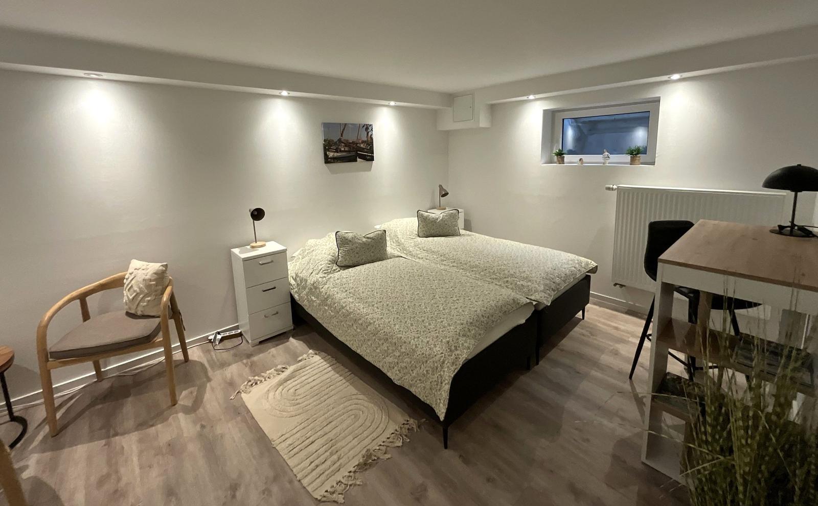 Schlafzimmer mit Bett, Nachttisch, Stuhl und Schreibtisch. Holzboden und Fenster.