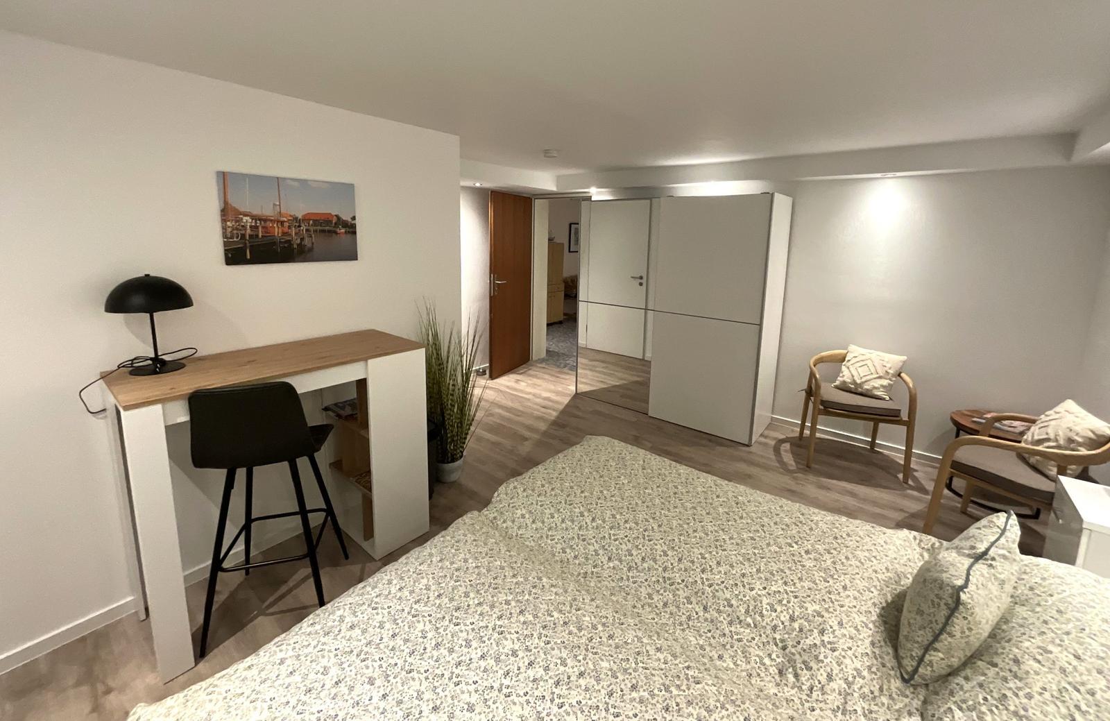 Schlafzimmer mit Schreibtisch, Stuhl und Bett. Hintergrund: Kleine Sitzgelegenheit und Schrank.