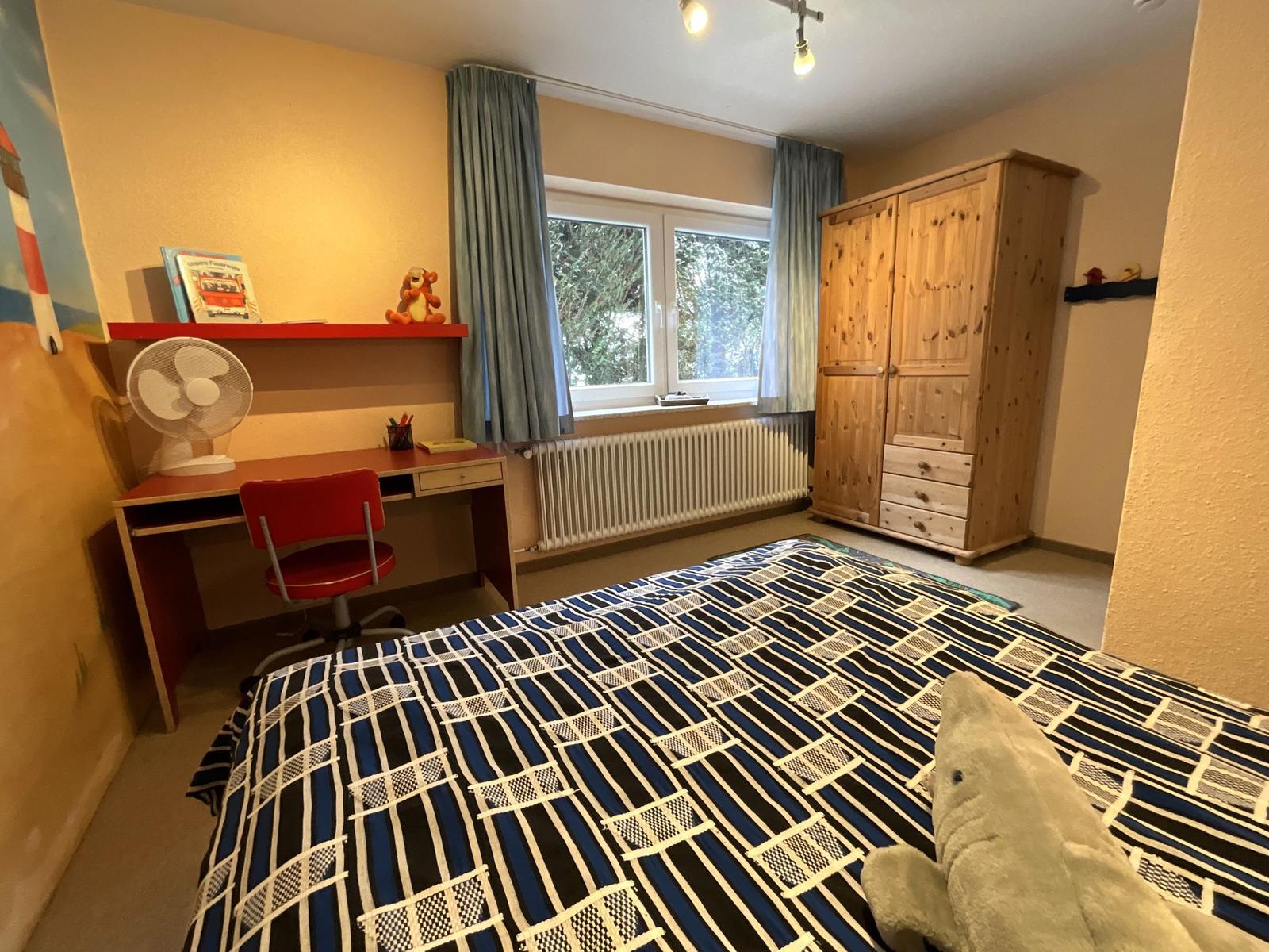 Schlafzimmer mit Bett, Schreibtisch und Holzschrank. Fenster mit Vorhängen.