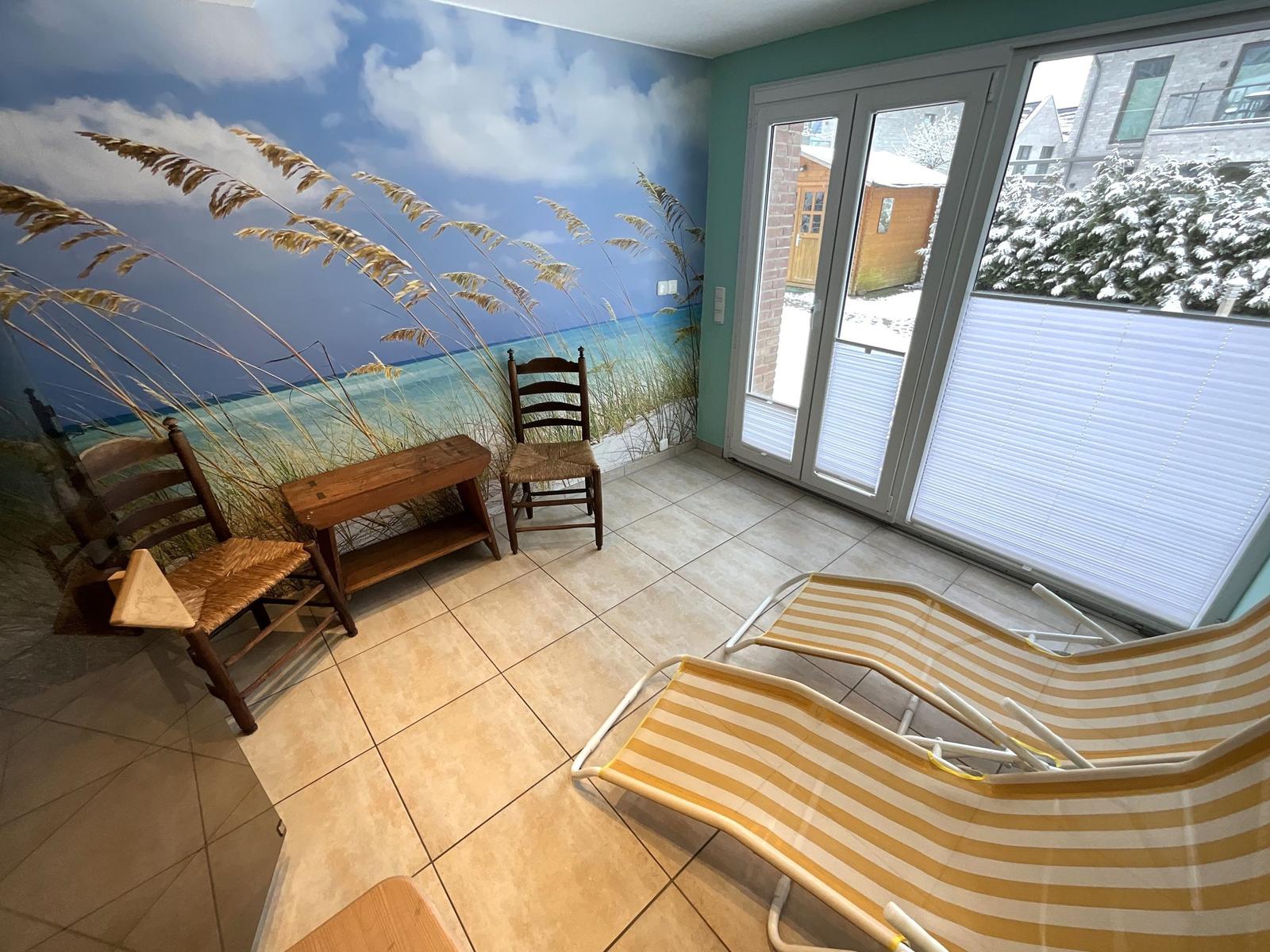 Raum mit Strandmotivwand, zwei Stühlen, Tisch und Liegestühlen. Durchgang zur Terrasse mit Blick auf Schnee.