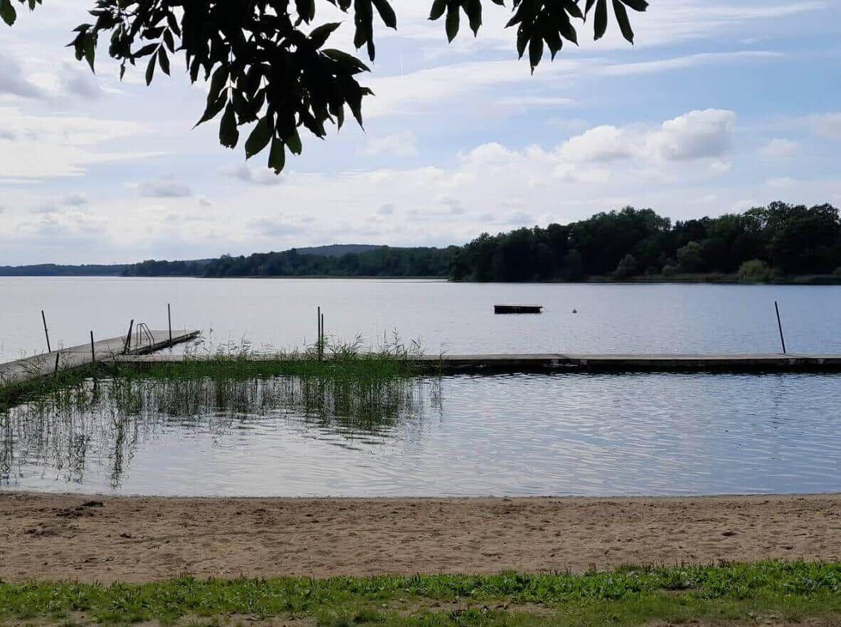 Strand mit Bootssteg und ruhigem See im Hintergrund.