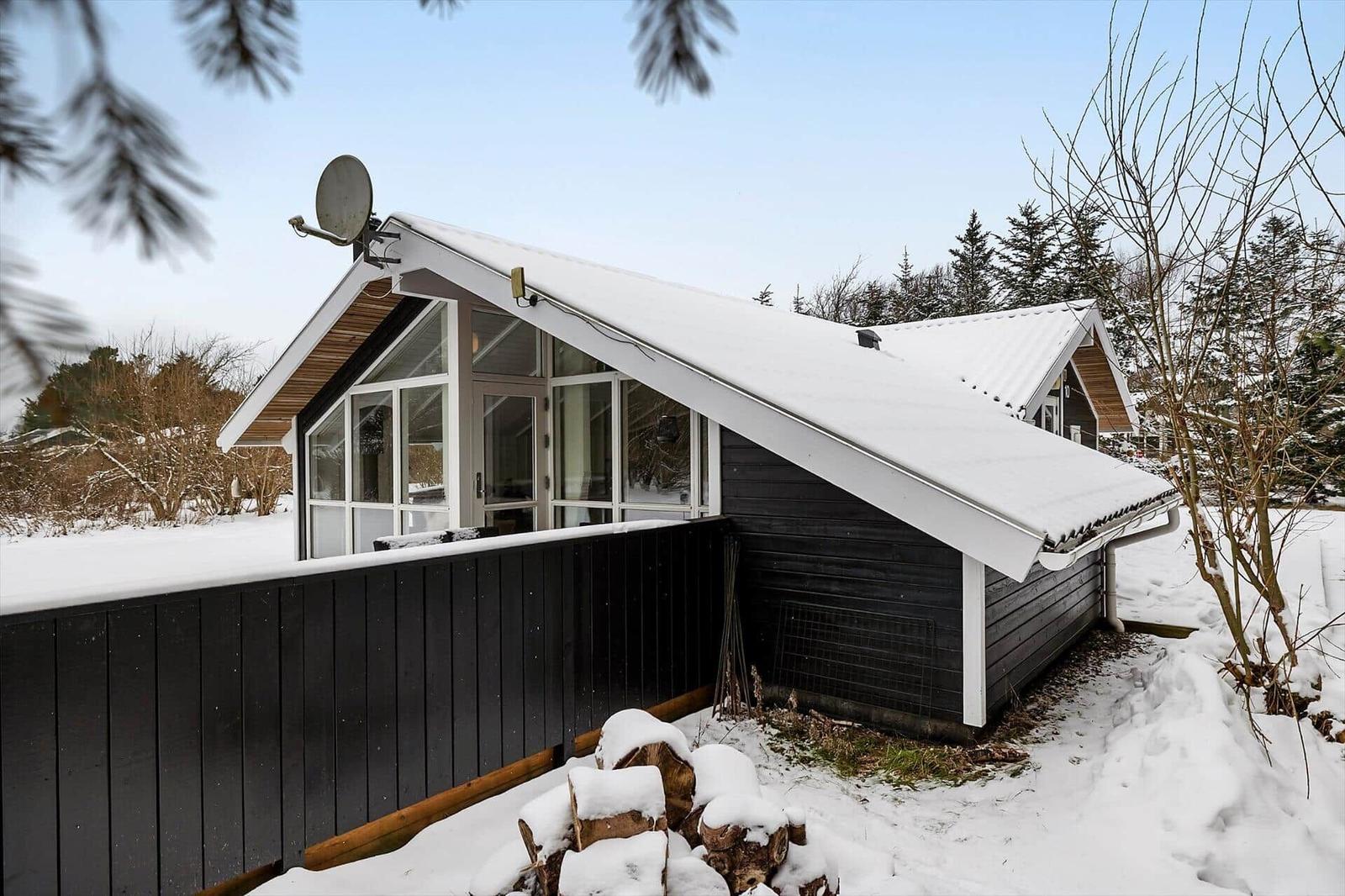 Black wooden house with snow, satellite dish, and woodpile in foreground.