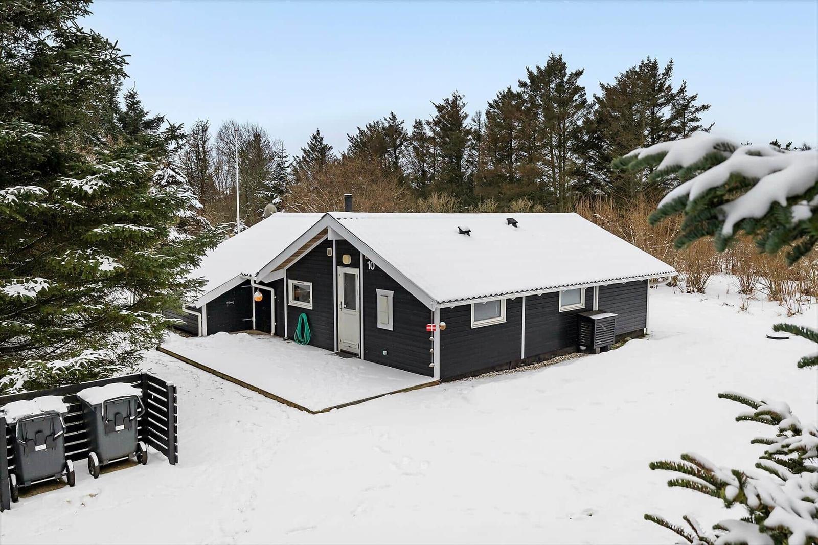 Black wooden house with snow, surrounded by evergreens and a snowy terrace.