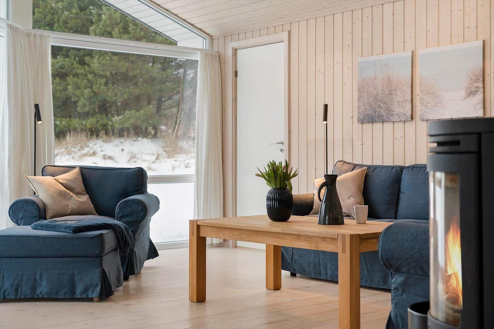Living room with sofa, coffee table, and fireplace. Large windows show forest and snow.
