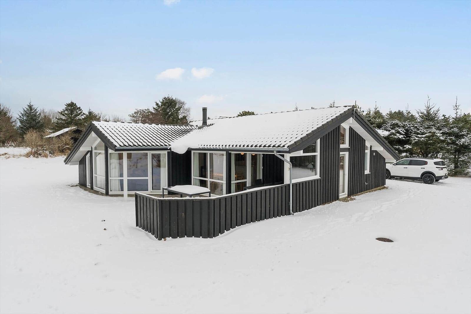 Black wooden house with snow, large window area, and white car in front.