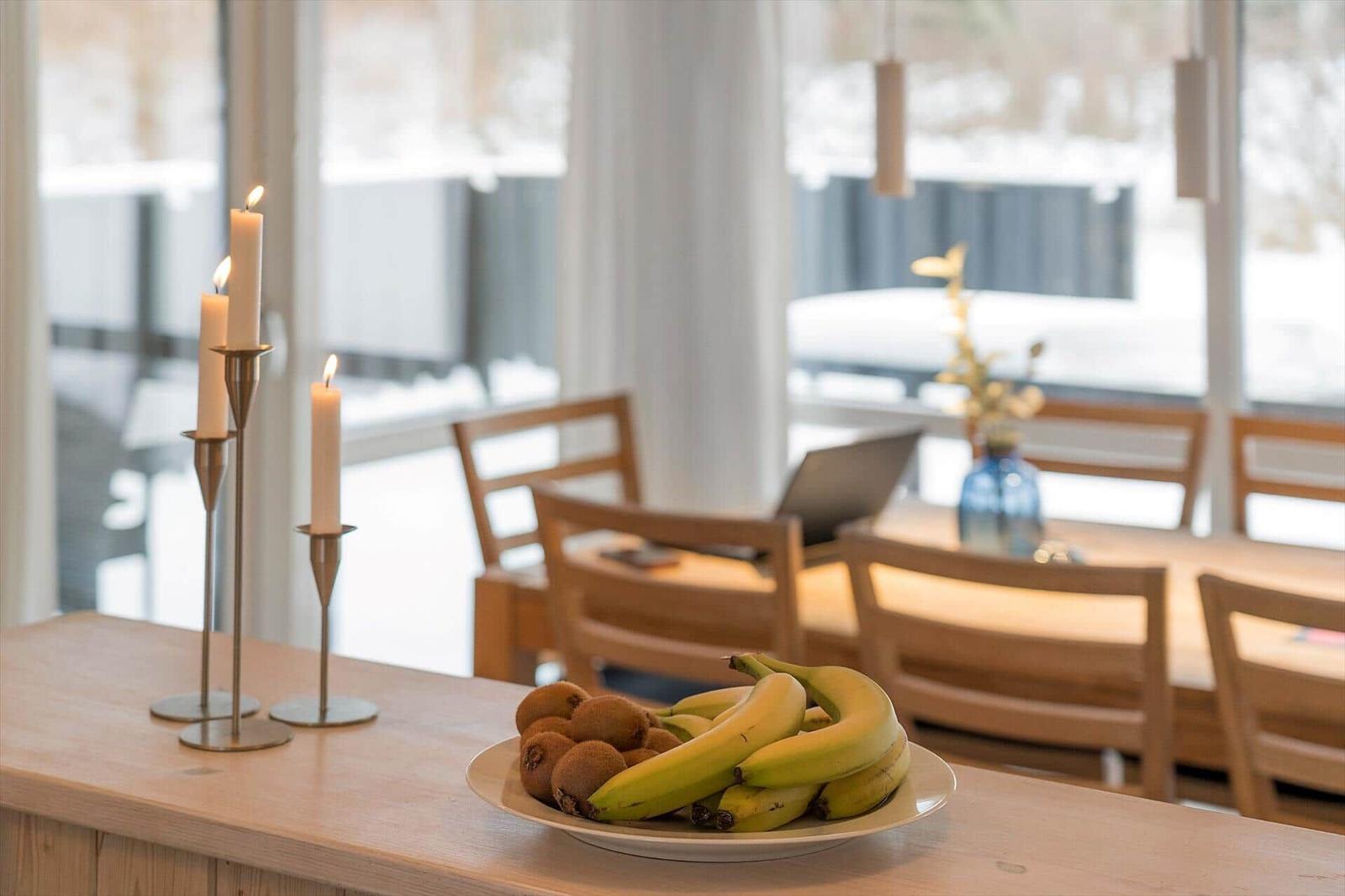 Fruit bowl and lit candles on kitchen island, dining area in background with snowy view.