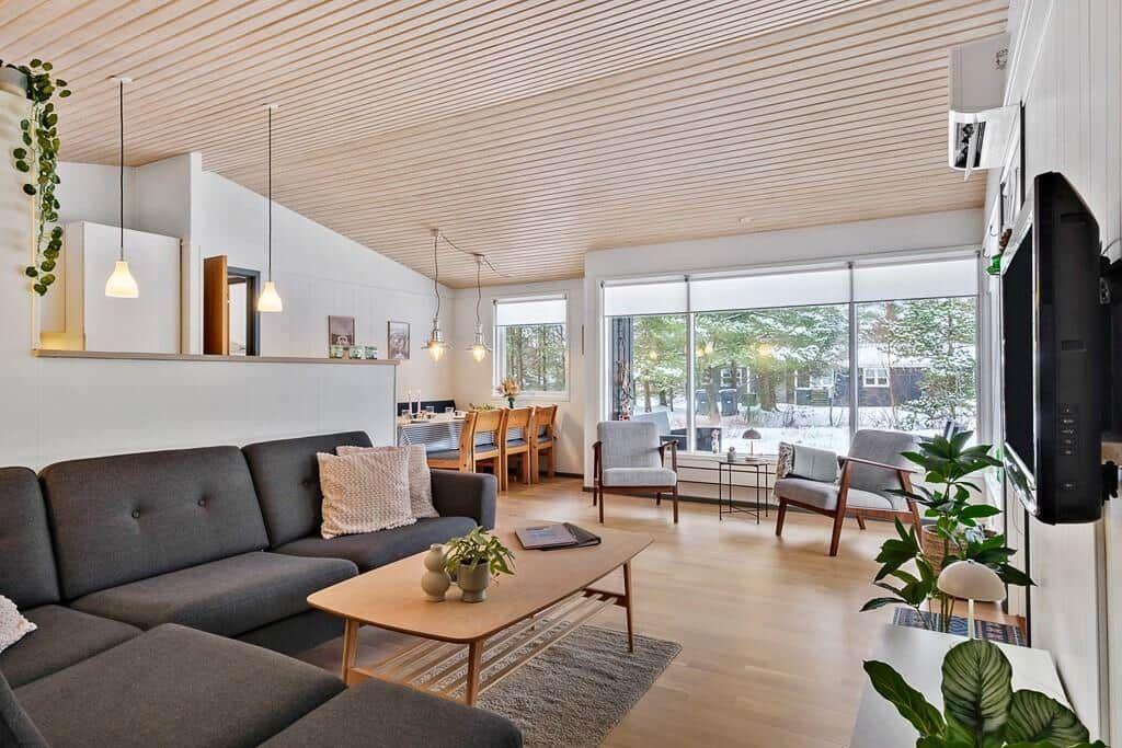 Living and dining area with sofa, table, and chairs. Large windows and wooden ceiling.