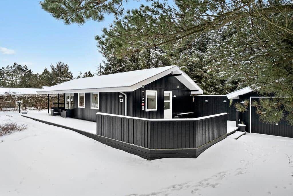 Black wooden cabin with snow-covered roof and terrace under trees.