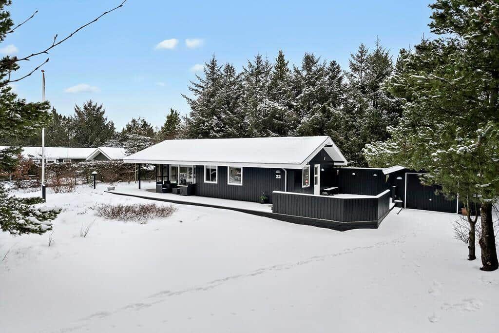 Black house with snow-covered roof and terrace among trees.