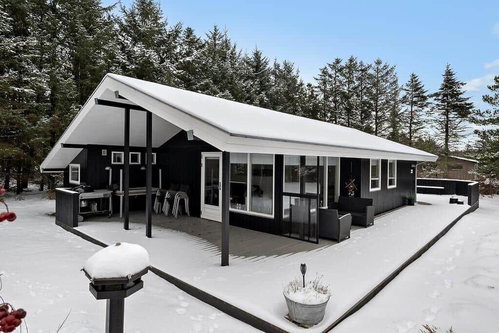 Black house with snow-covered roof and patio in the woods