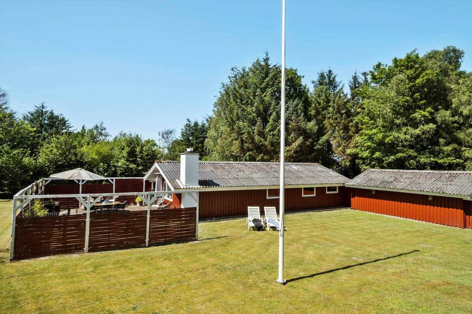Red wooden house with terrace, garden, and lounge chairs. Background: dense trees.