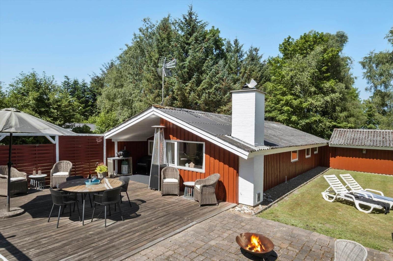 Red vacation house with terrace, grill, and lawn area in green surroundings.