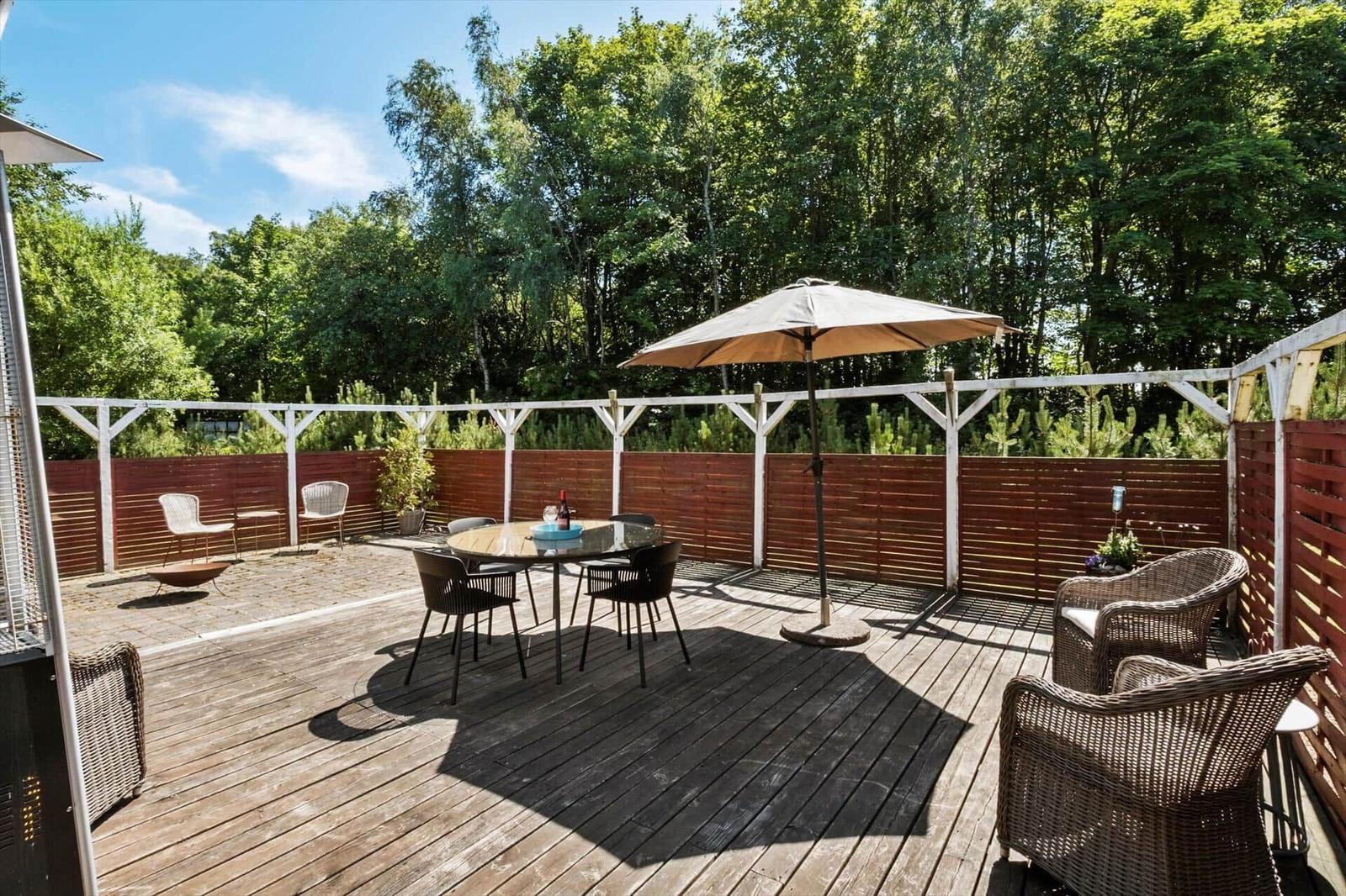 Terrace with table, chairs, and umbrella. Surrounded by forest and wooden railing.