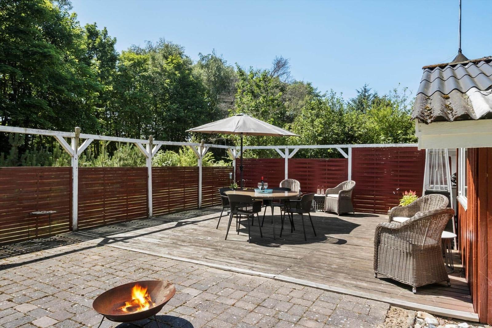 Terrace with seating, umbrella, and fire pit. Surrounded by wooden fence and trees.