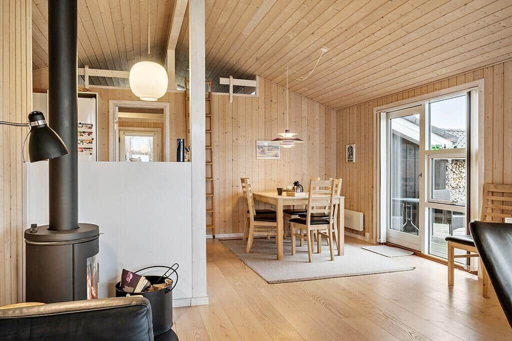 Wood-paneled kitchen with dining table, chairs, and windows. Wood-burning stove and seating area in foreground.