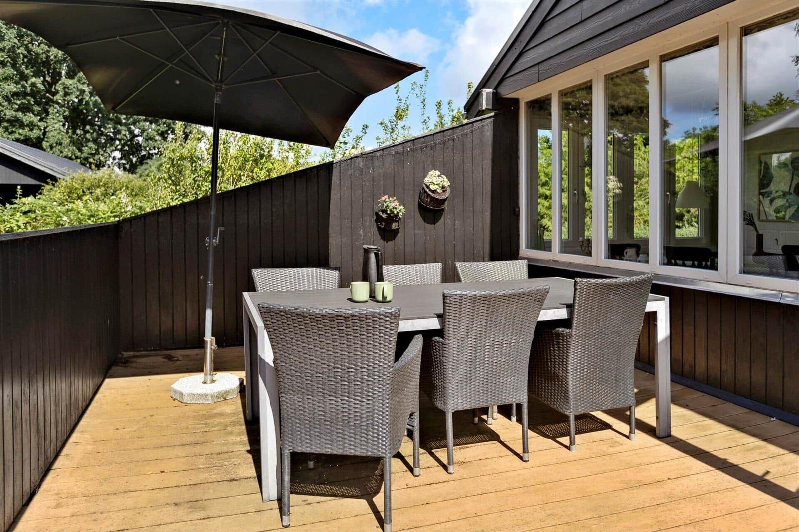 Terrace with table, six chairs, and sun umbrella. Wooden deck and dark fence.