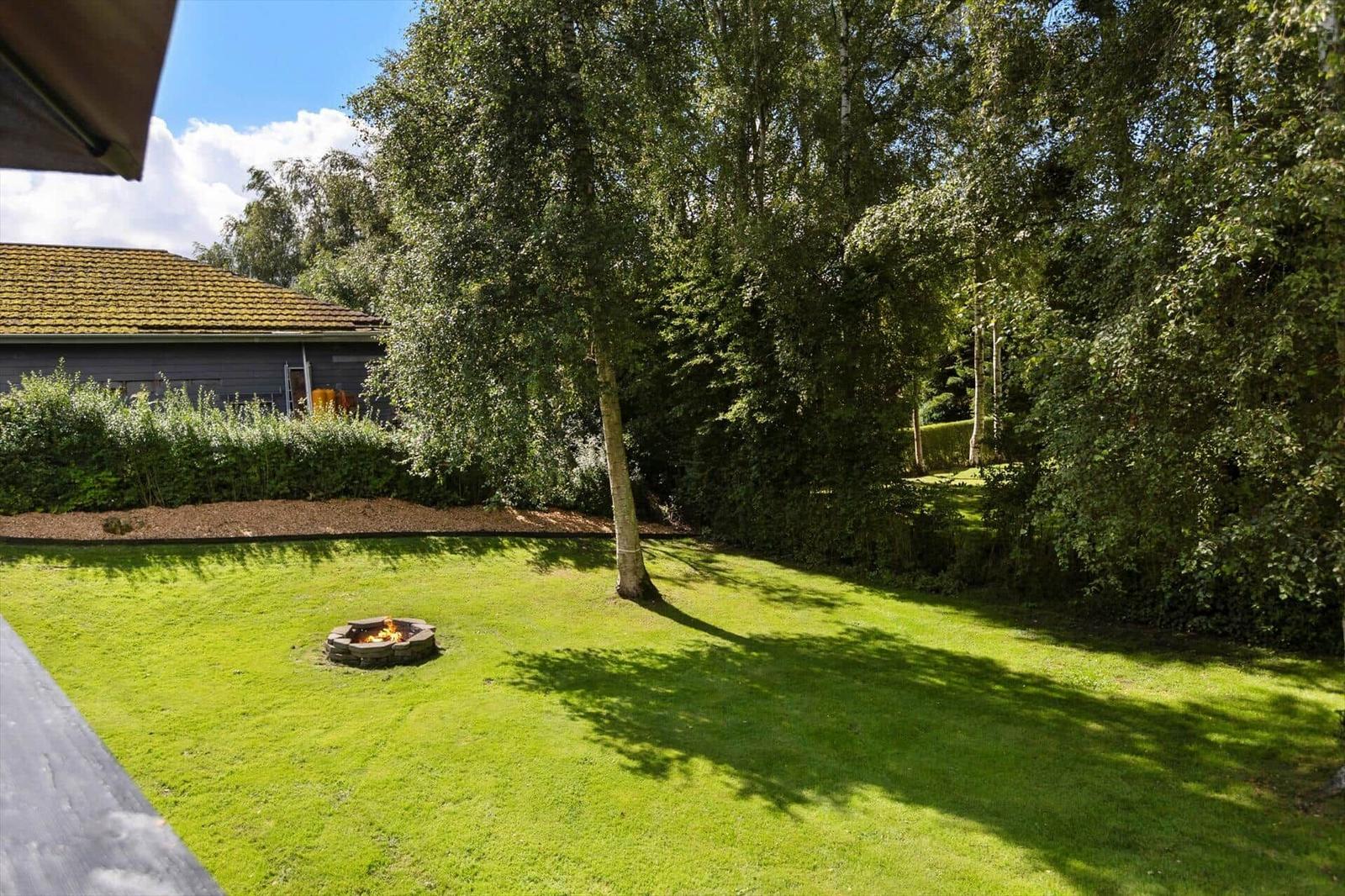 Green garden with fire pit and trees. House with roof in background.