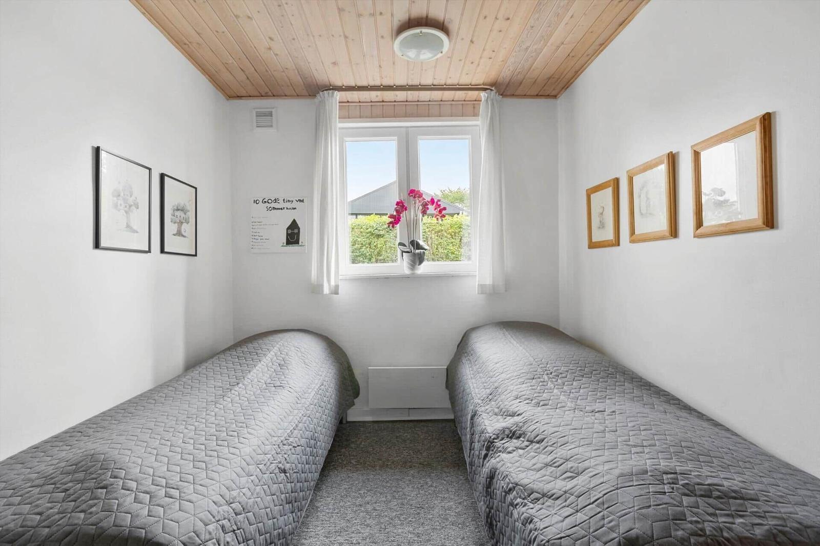 Two beds with gray quilt, wooden ceiling, and window with flowers.