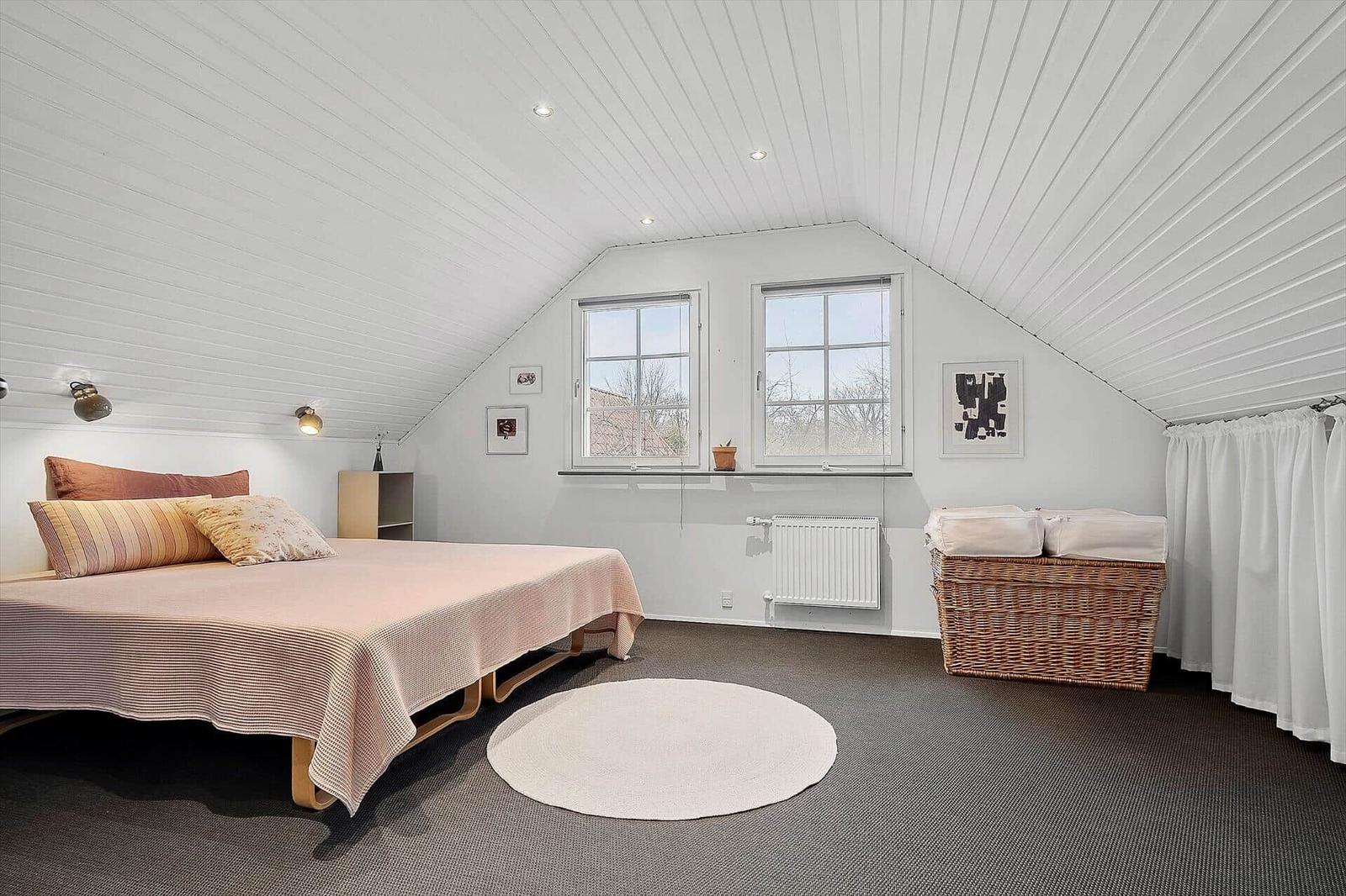 Schlafzimmer mit Dachschrägen, Bett, zwei Fenstern und Korb.