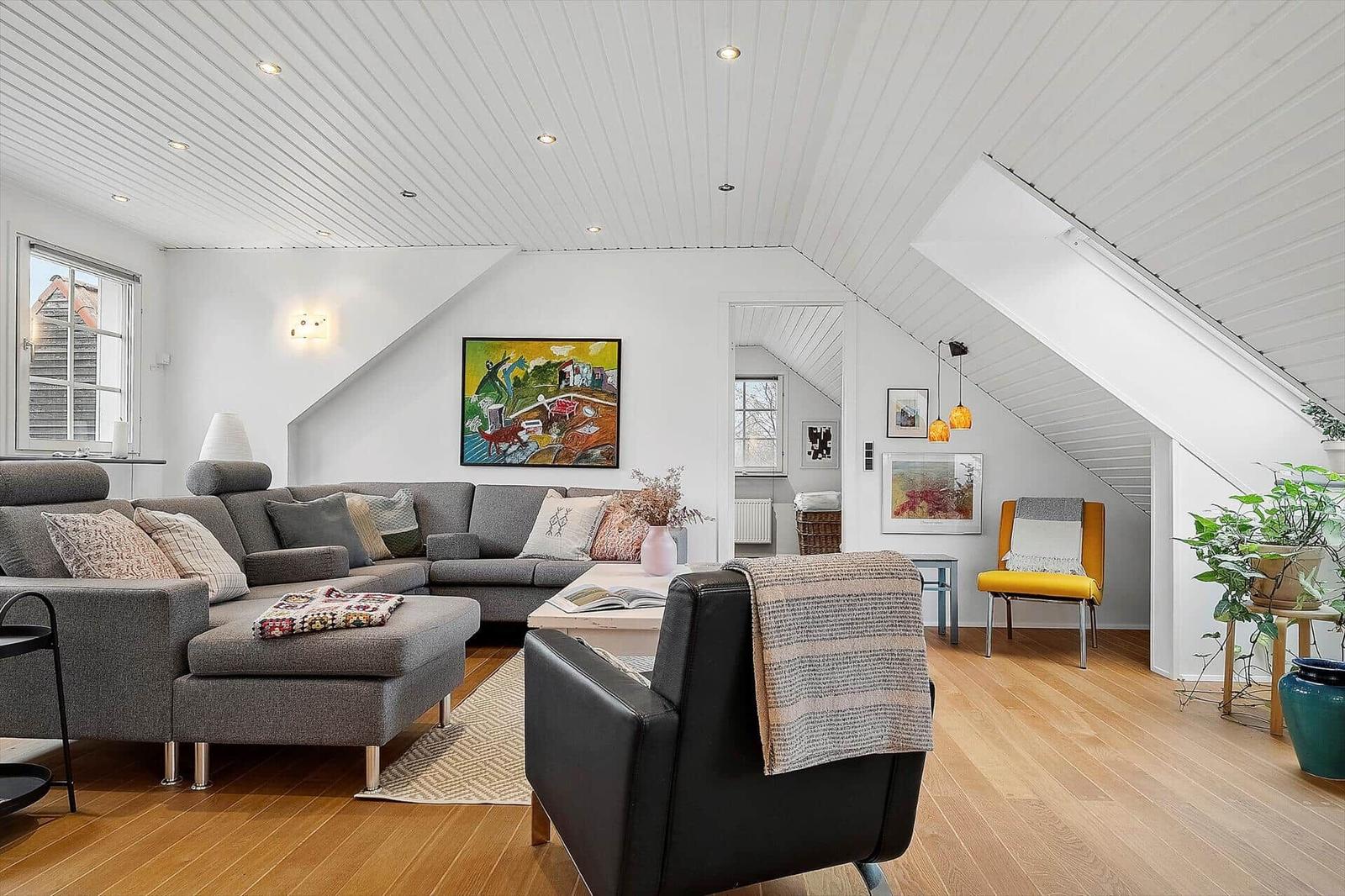 Wohnzimmer mit grauem Sofa, schwarzer Ledercouch und Holzboden unter schrägen weißen Wänden.