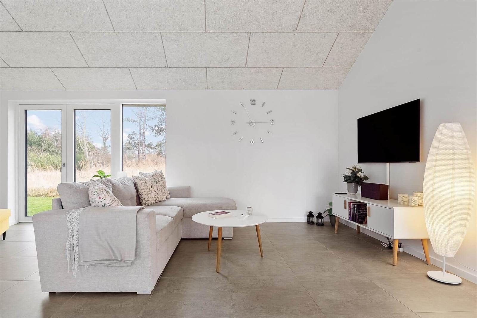Wohnzimmer mit Sofa, Tisch und Fernseher. Große Fenster mit Blick auf Grünflächen.