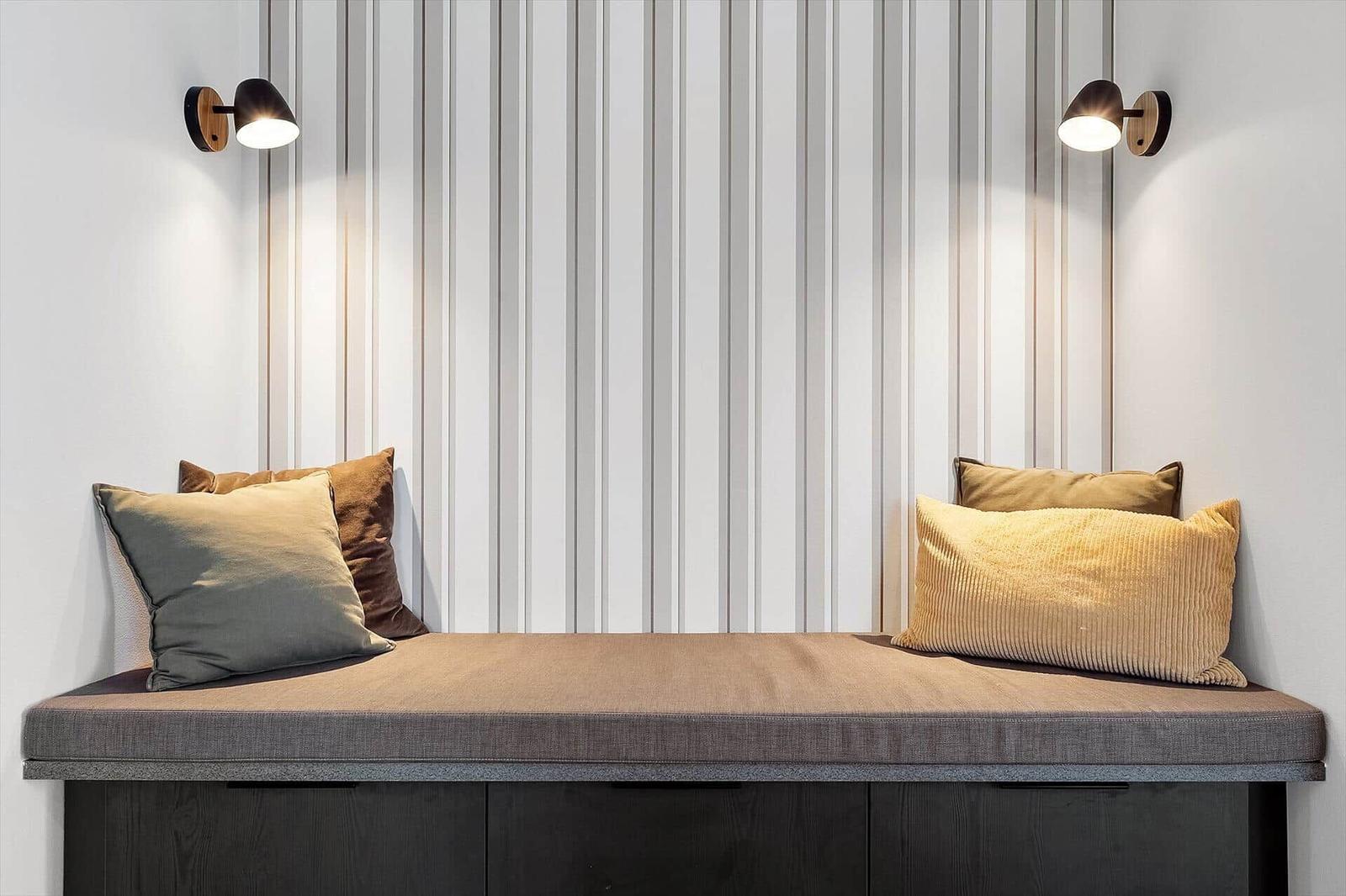 Sitzbank mit Kissen und Wandlampen an gestreifter Wand.