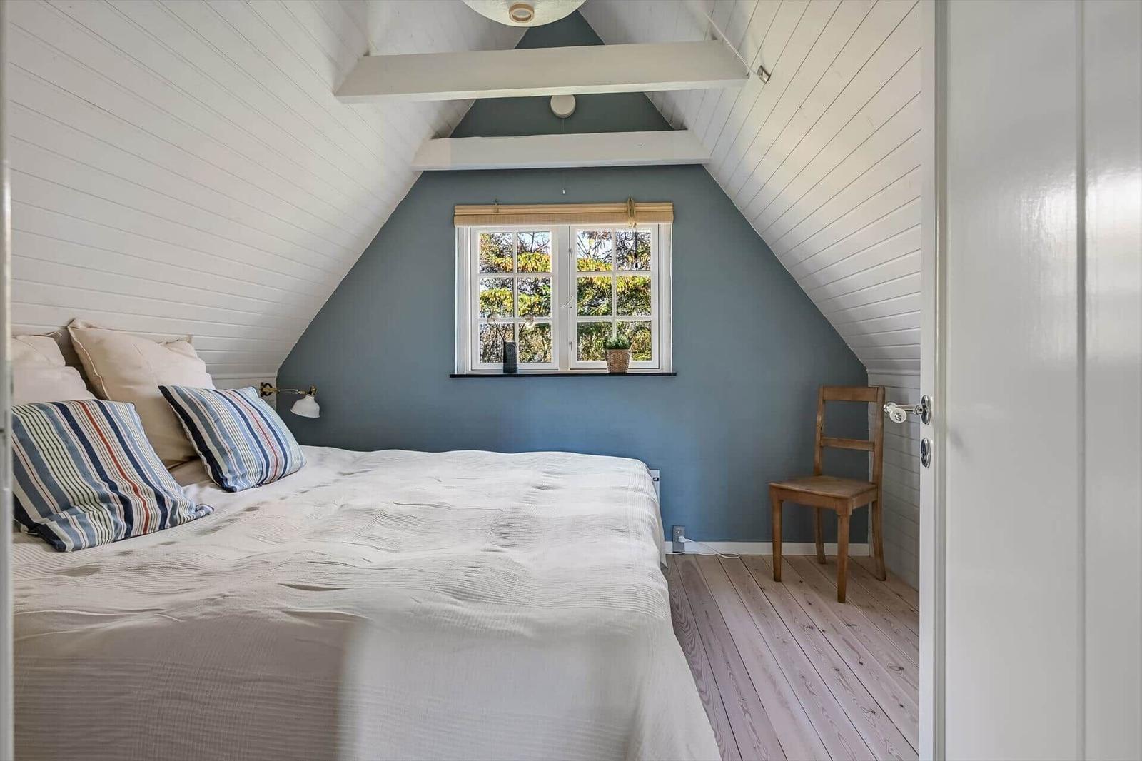 Schlafzimmer mit Dachschrägen, Bett, Stuhl und Fenster mit Blick auf Bäume.