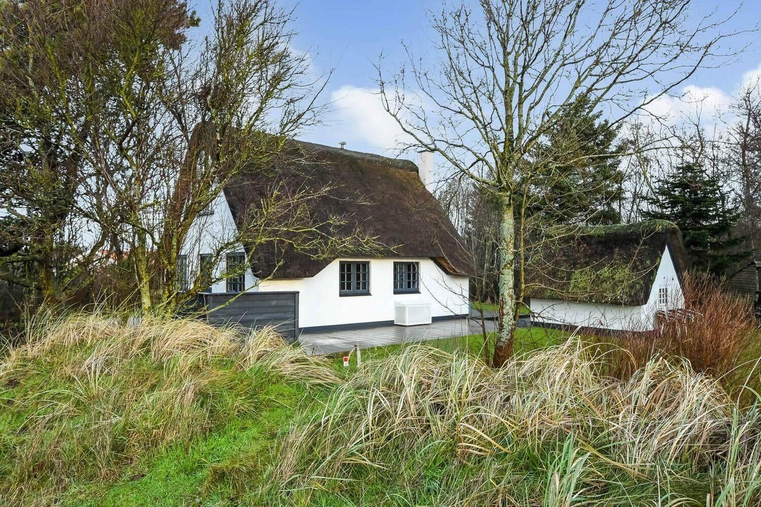 Weiße Hütte mit strohgedecktem Dach, umgeben von Bäumen und Gras.