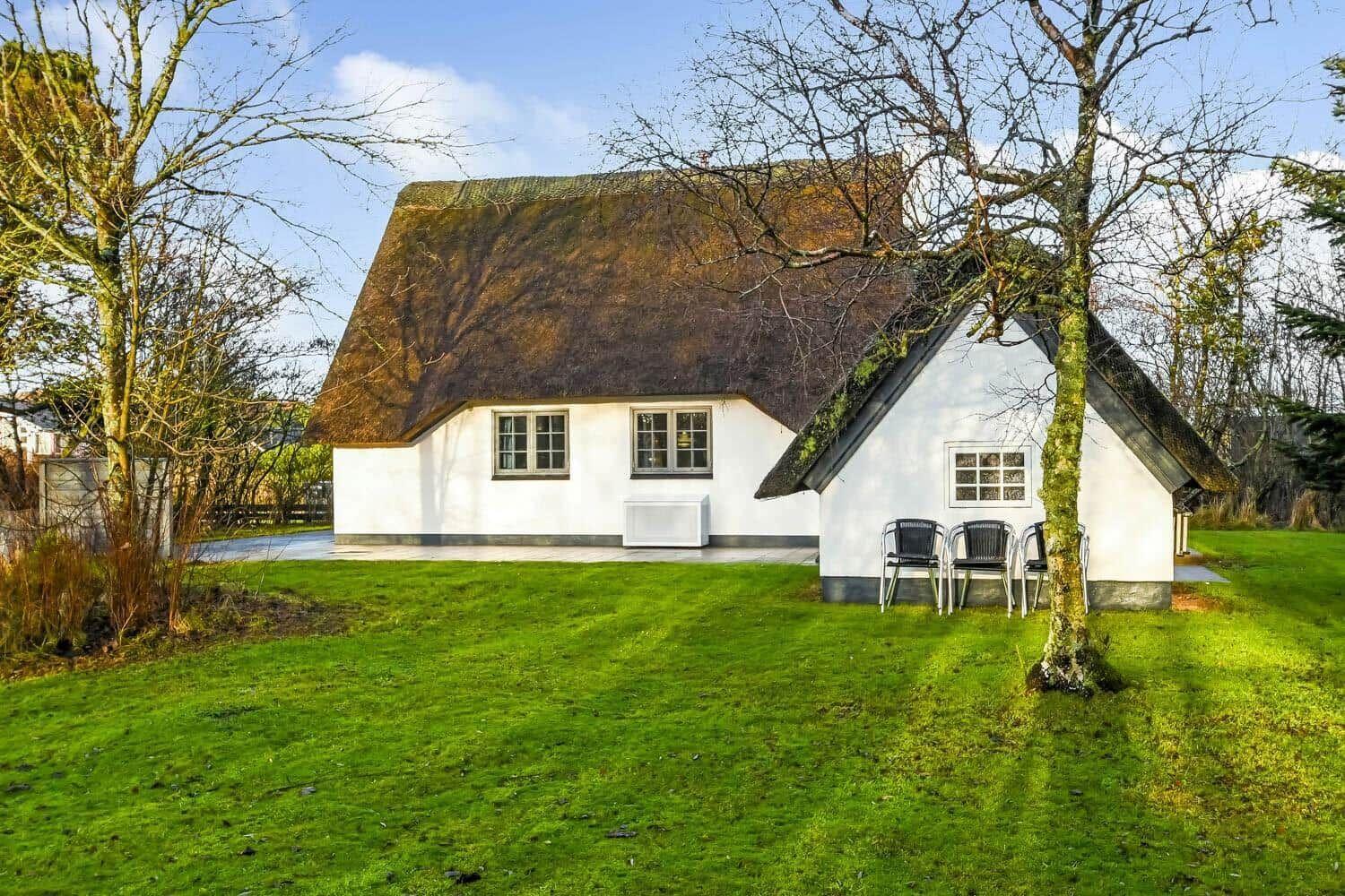 Wohnhaus mit Strohdach und grünem Rasen. Außentisch mit Stühlen unter Bäumen.