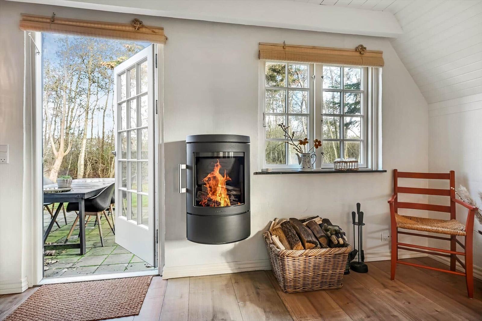 Raum mit Kamin, Fenster und Tür zum Garten. Holzstuhl und Korb mit Brennholz.