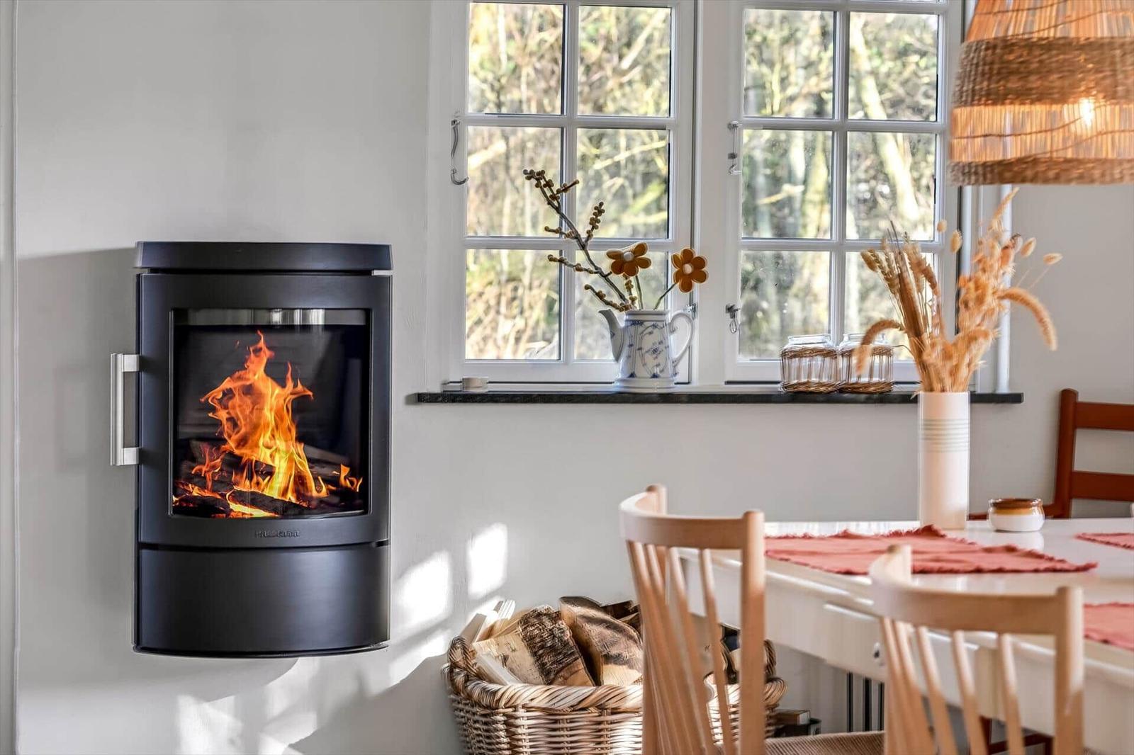Wohnraum mit Kaminofen, Tisch und Stühlen, Fenster mit Blumen und Licht.