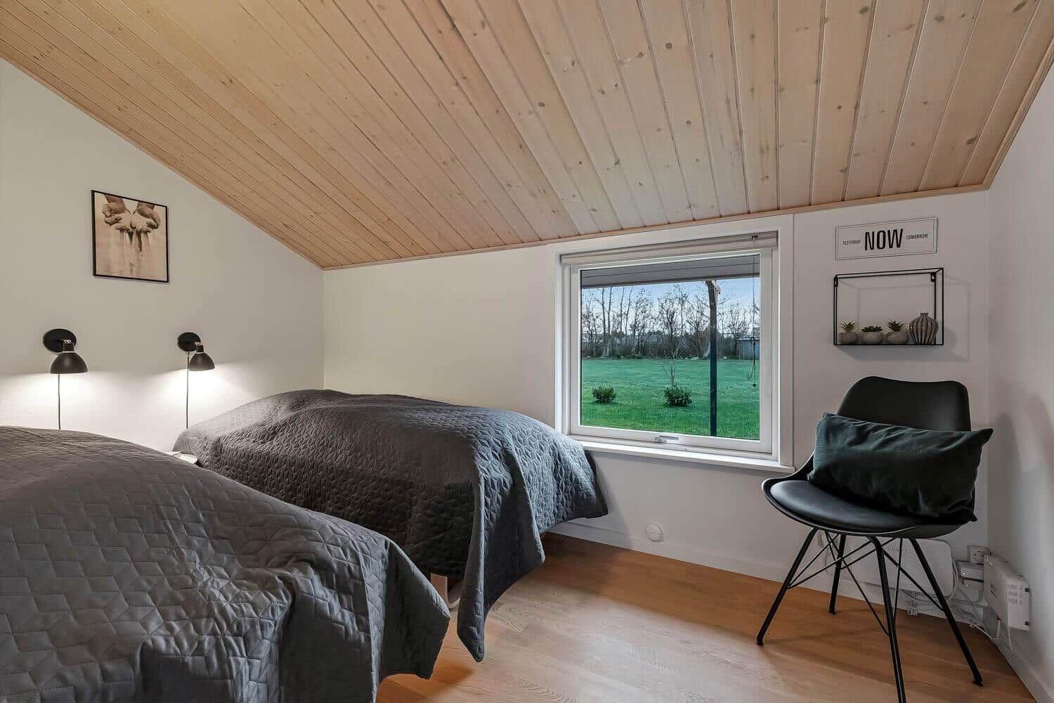 Zwei Betten mit grauen Bettdecken, Holzdecke, Fenster mit Blick auf Grünfläche und schwarze Sitzgelegenheit.