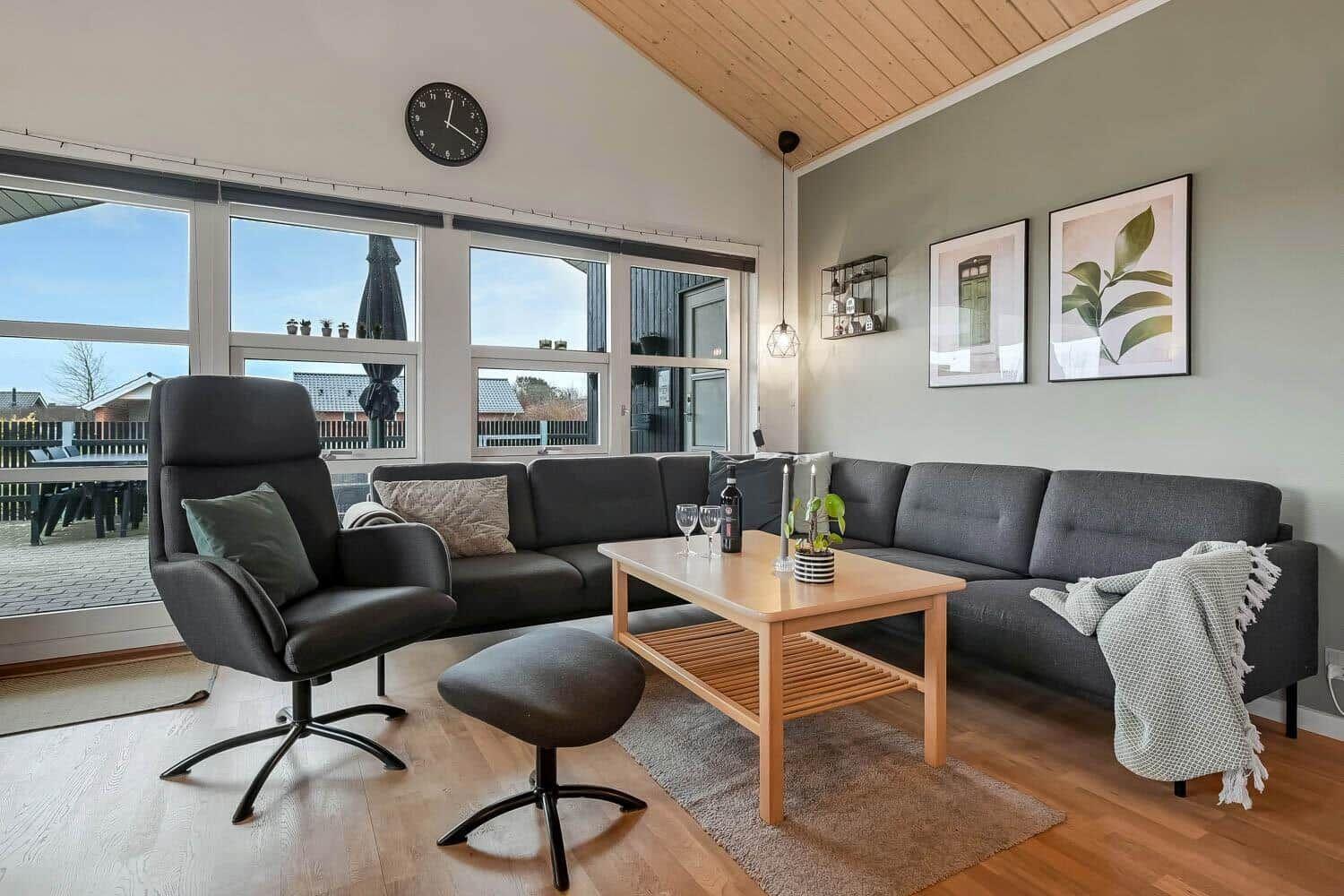 Wohnzimmer mit Sofa, Sessel und Tisch. Große Fenster und Holzdecke.
